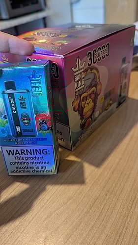 Deux boîtes de produits de vapotage aux couleurs vives sur une table, comportant une grande étiquette d'avertissement sur la dépendance à la nicotine.