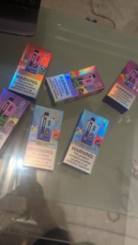 Várias caixas de dispositivos vape descartáveis coloridos rotulados como "Bang King" são exibidas em uma superfície de vidro. A embalagem apresenta várias imagens de sabores de frutas e avisos proeminentes sobre o vício em nicotina.